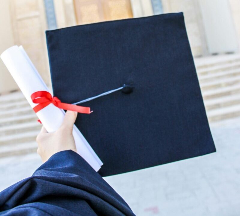 A hand holding a graduation hat and diploma in front of a university building.